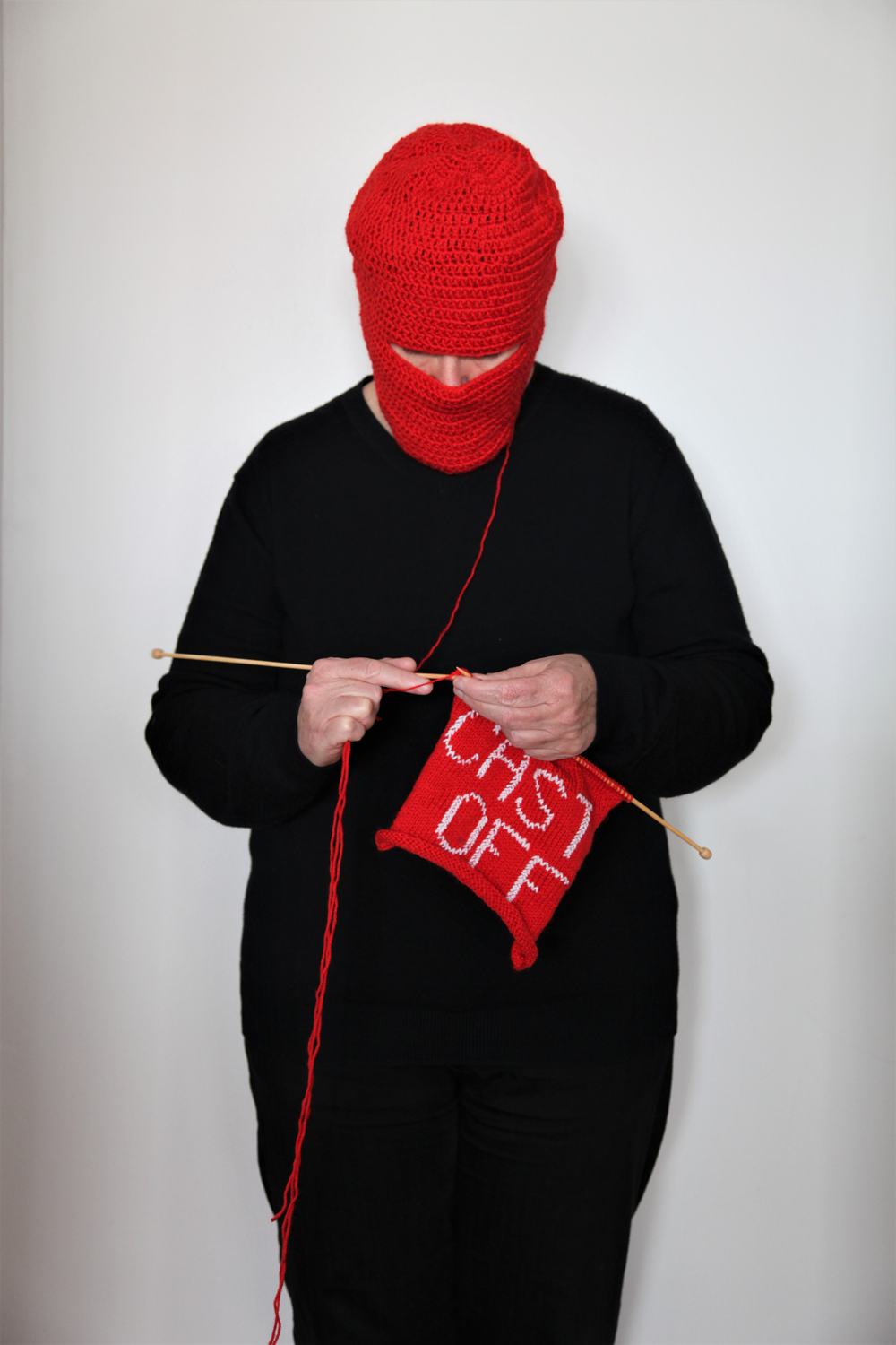 Person wearing and unravelling a red crochet mask, knitting the unraveled red wool into a panel with text in white saying cast off. 