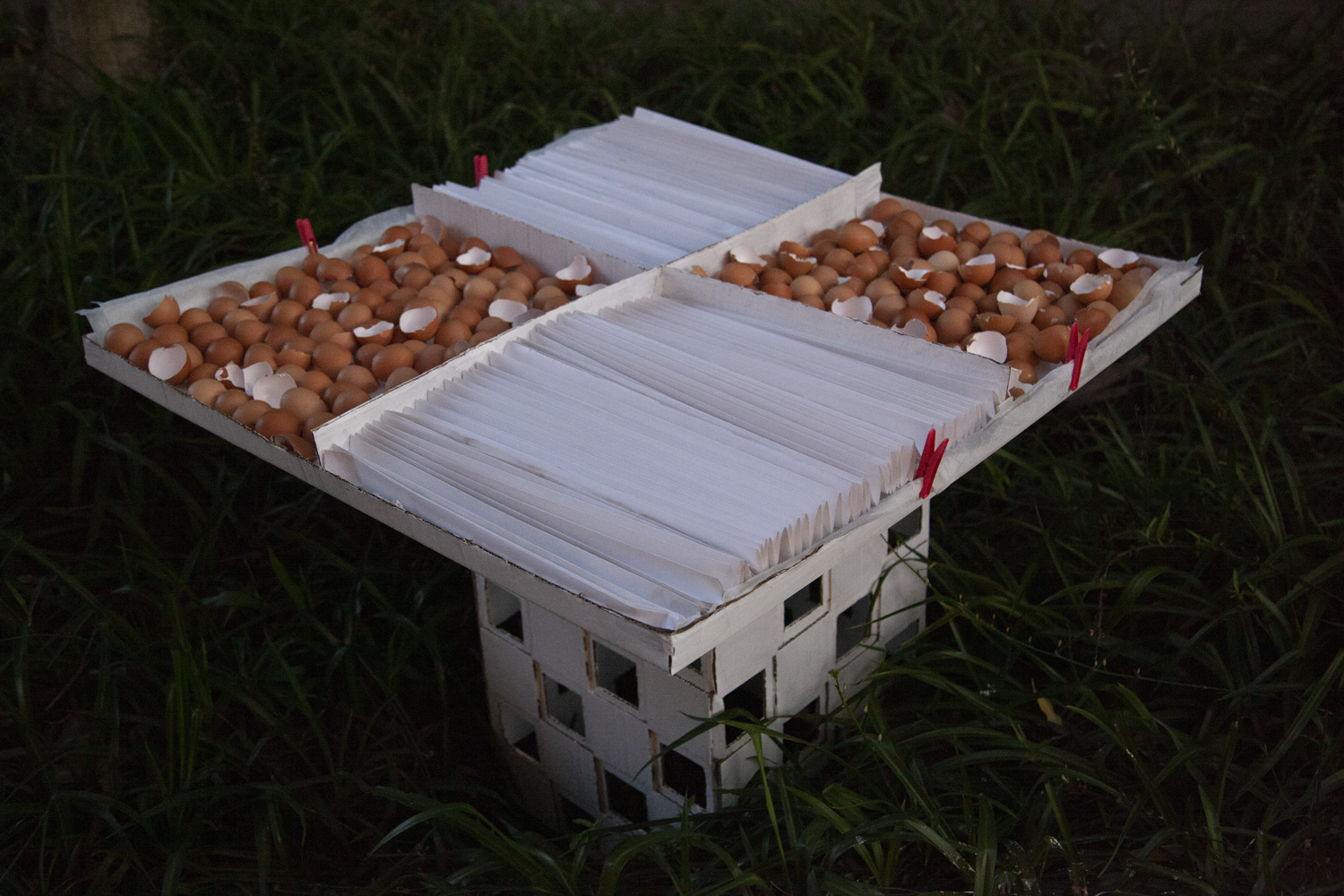 Painted white cardboard monument holding extended flat tray. Tray lined with baking paper and contains eggshells and accordian-folded paper across four alternating square sections.