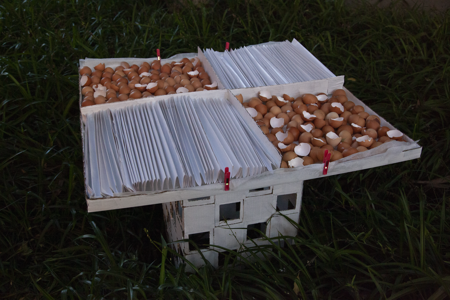 Painted white cardboard monument holding extended flat tray. Tray lined with baking paper and contains eggshells and accordian-folded paper across four alternating square sections.