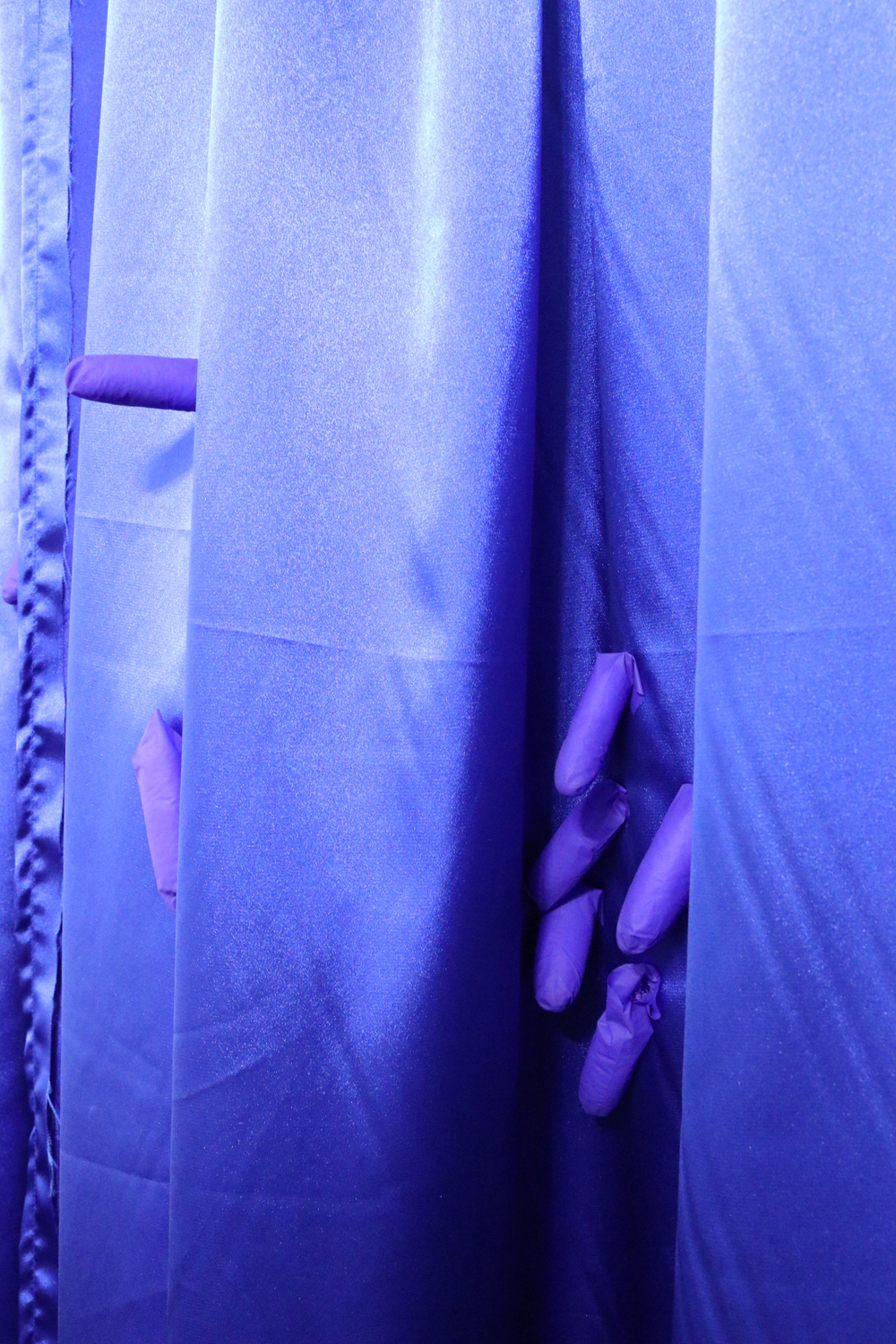 A close-up of blue satin curtains. There are blue latex appendages clustered in the folds of the curtain. 