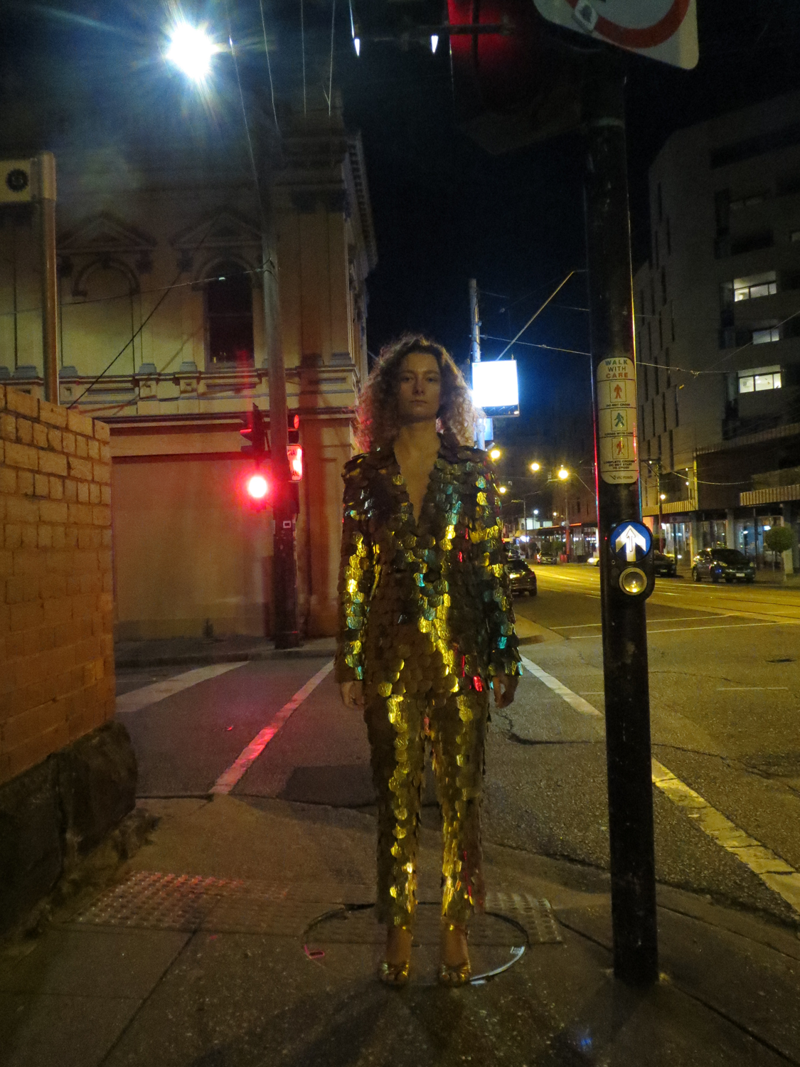 Translucent dissolving suit with red stitching being washed off body by water. Gold scaly sequin covered suit, reflecting daylight and traffic lights.