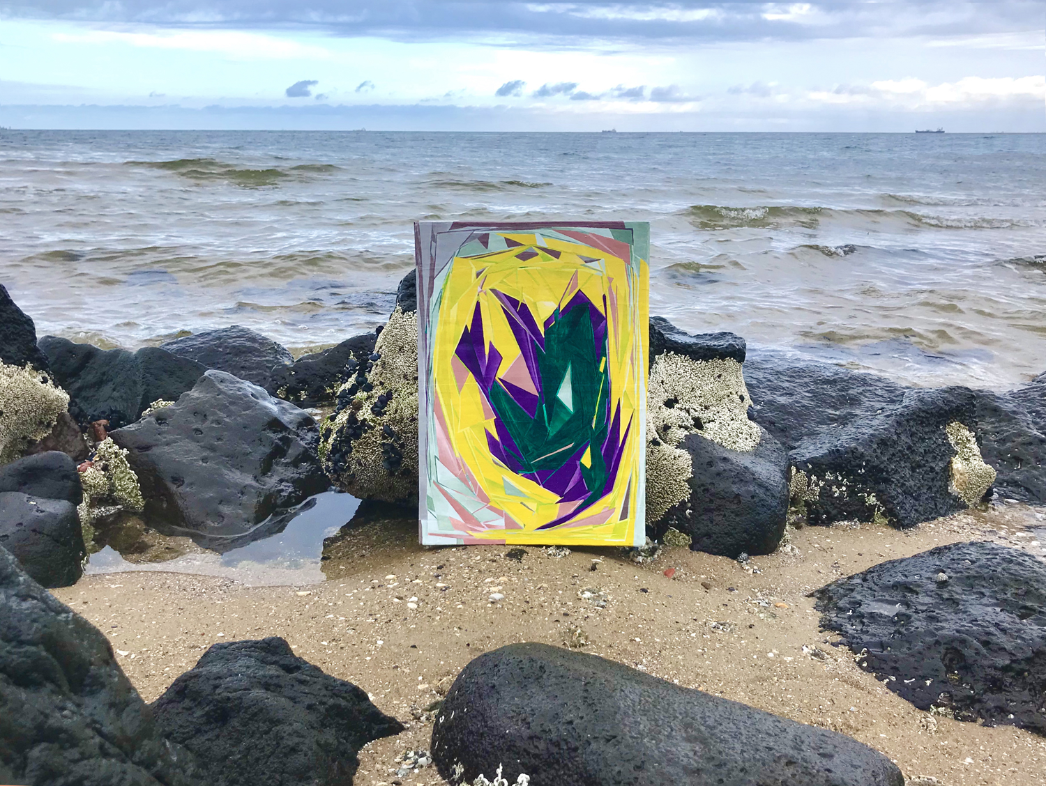 Digital photograph of the installation of the painting titled "An Uncaused Joy" situated amongst rocks on the sea shore.