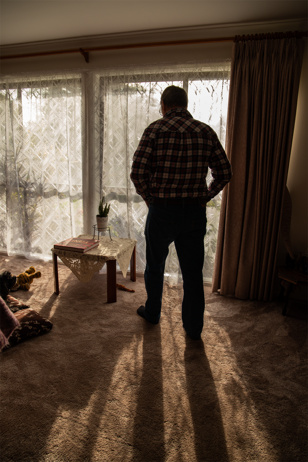 Digital photograph of a man standing in dark living room. His back is faced towards the camera, as he looks to the ground with his hands in pockets.