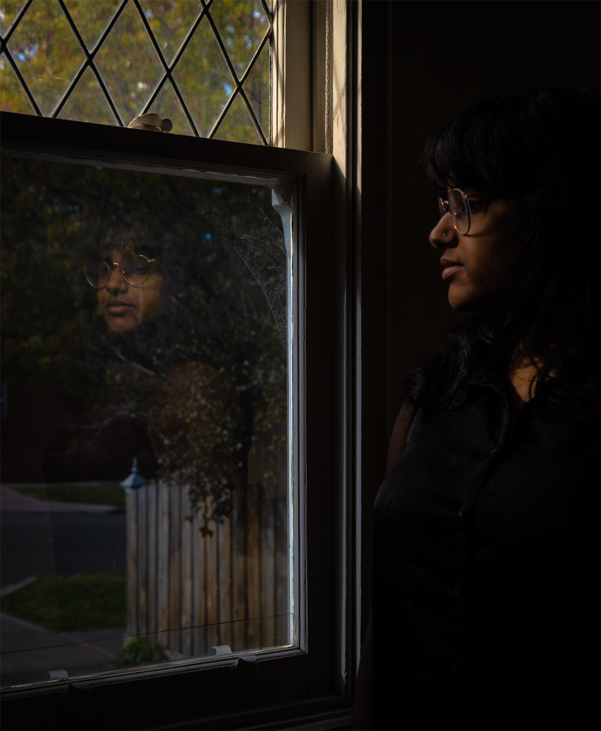 Digital photograph of a young woman looking out of the window of a darkened room. Her reflection can be seen in the light of the window pane.