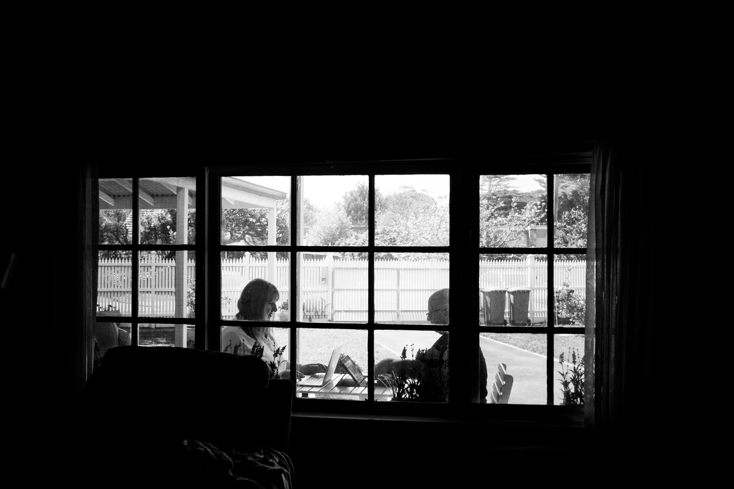 Black and white digital photograph of an interior window revealing a couple sitting outside at a table working on laptops.