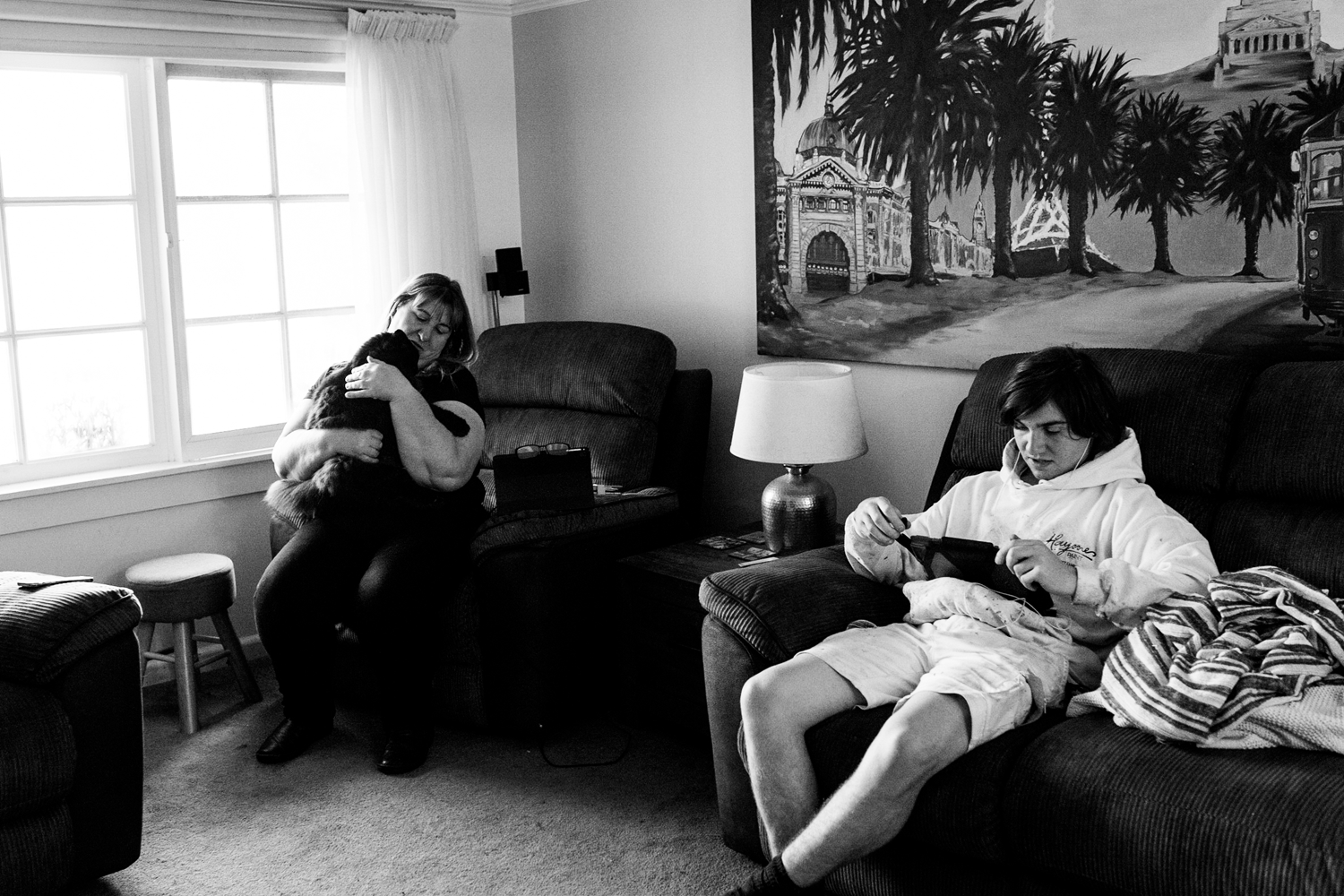 Black and white candid photograph of a woman and a teenage boy sitting in a residential lounge room. Woman holds dog. 