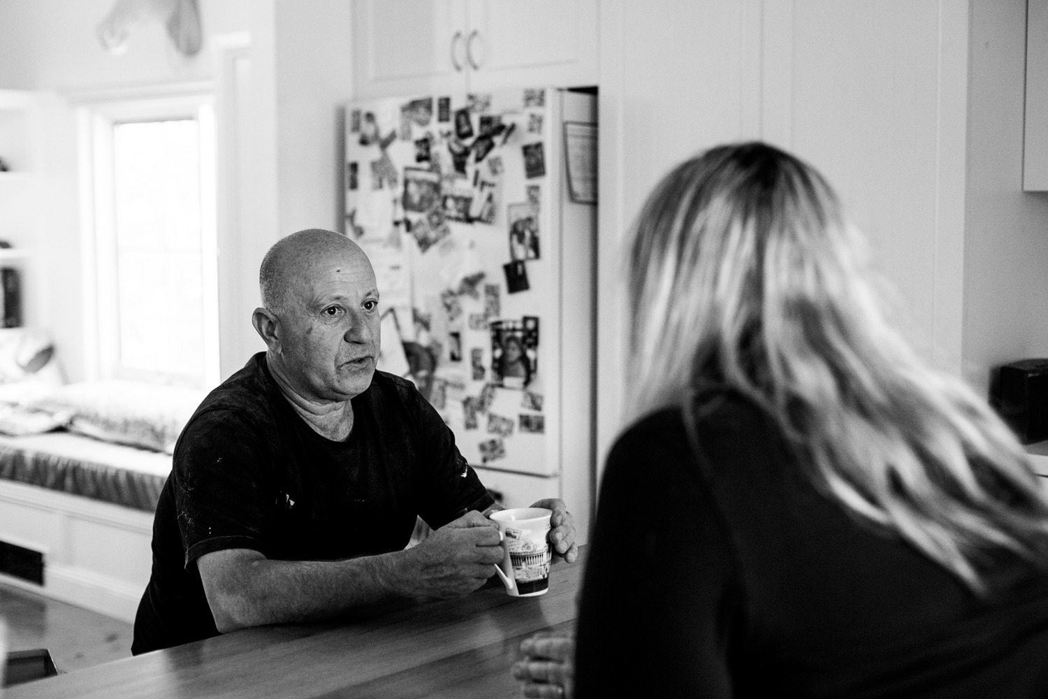 Black and white image of couple sitting at a kitchen bench in suburban home. Couple are in conversation with woman's back facing the camera.