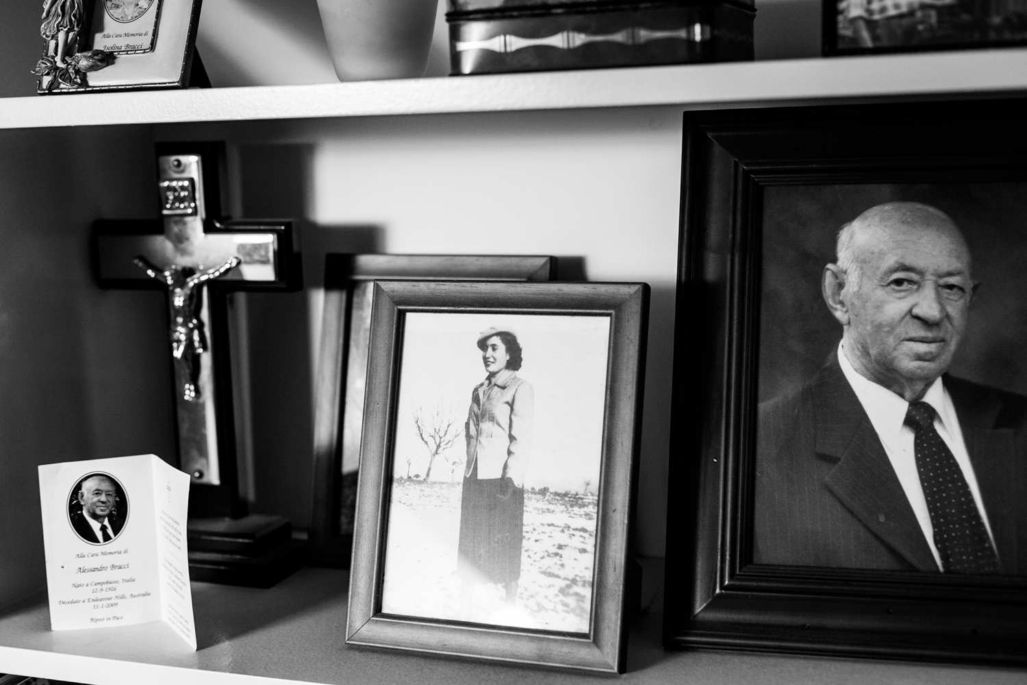 Black and white photograph of a bookshelf with a photo frame holding a photograph of woman, a funeral pamphlet, and a cross. 