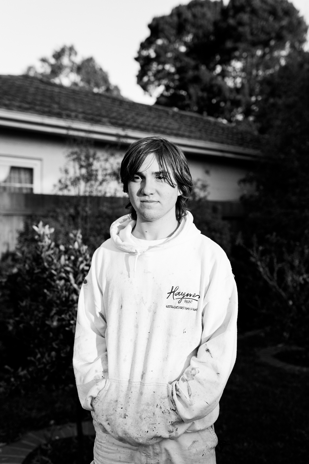 Black and white photograph of a portrait of a young man standing in suburban backyard wearing paint covered jumper.