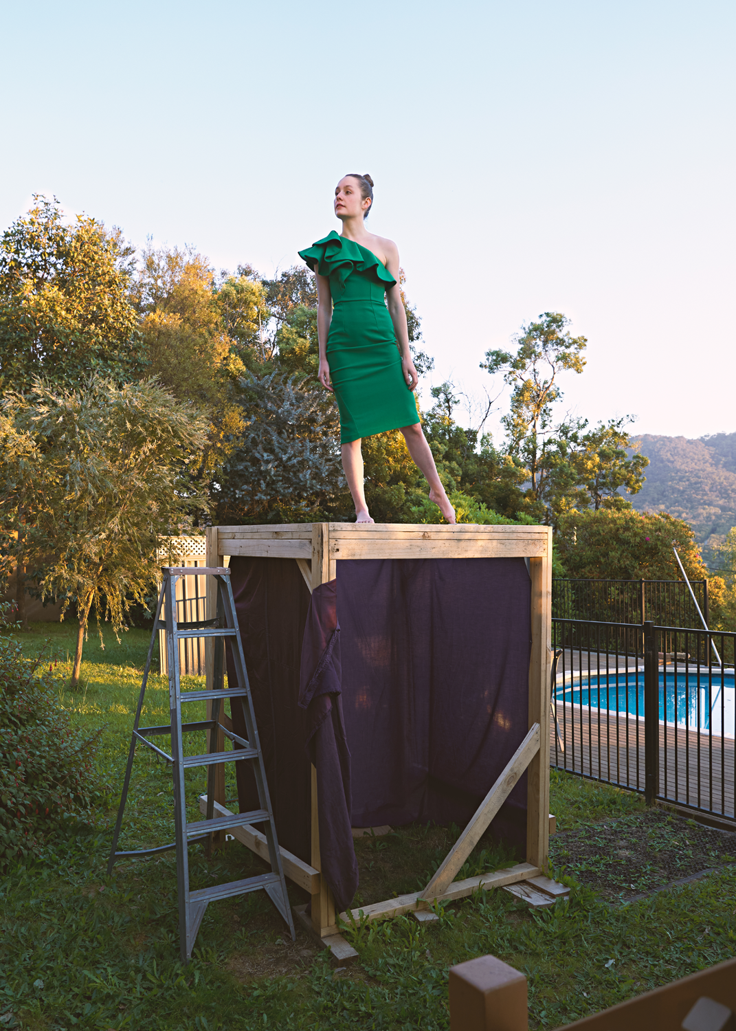 Colour video still of barefoot woman in green dress standing on perspex platform structure in suburban front yard.