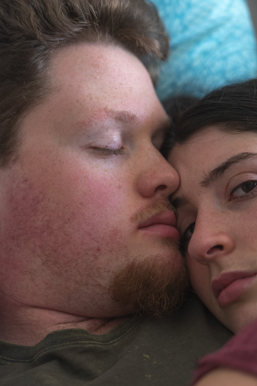 Colour photograph of extreme closeup of couple in bed, cuddling up, male asleep, female from before looking at camera.