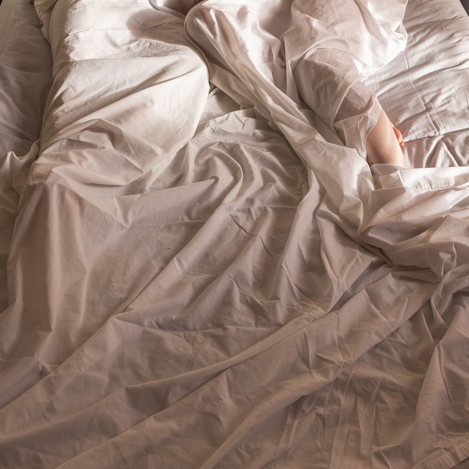 Colour photograph of closeup of bottom half of bed with messy sheets with two sets of legs under the bedsheets. 