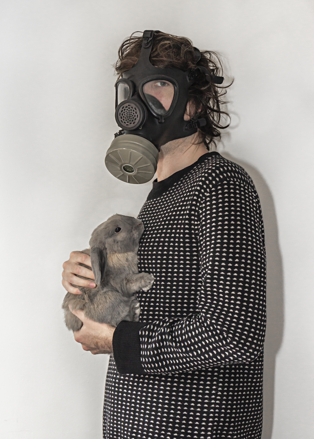 Colour photograph of man in black jumper holding a grey rabbit, wearing a black gas mask.