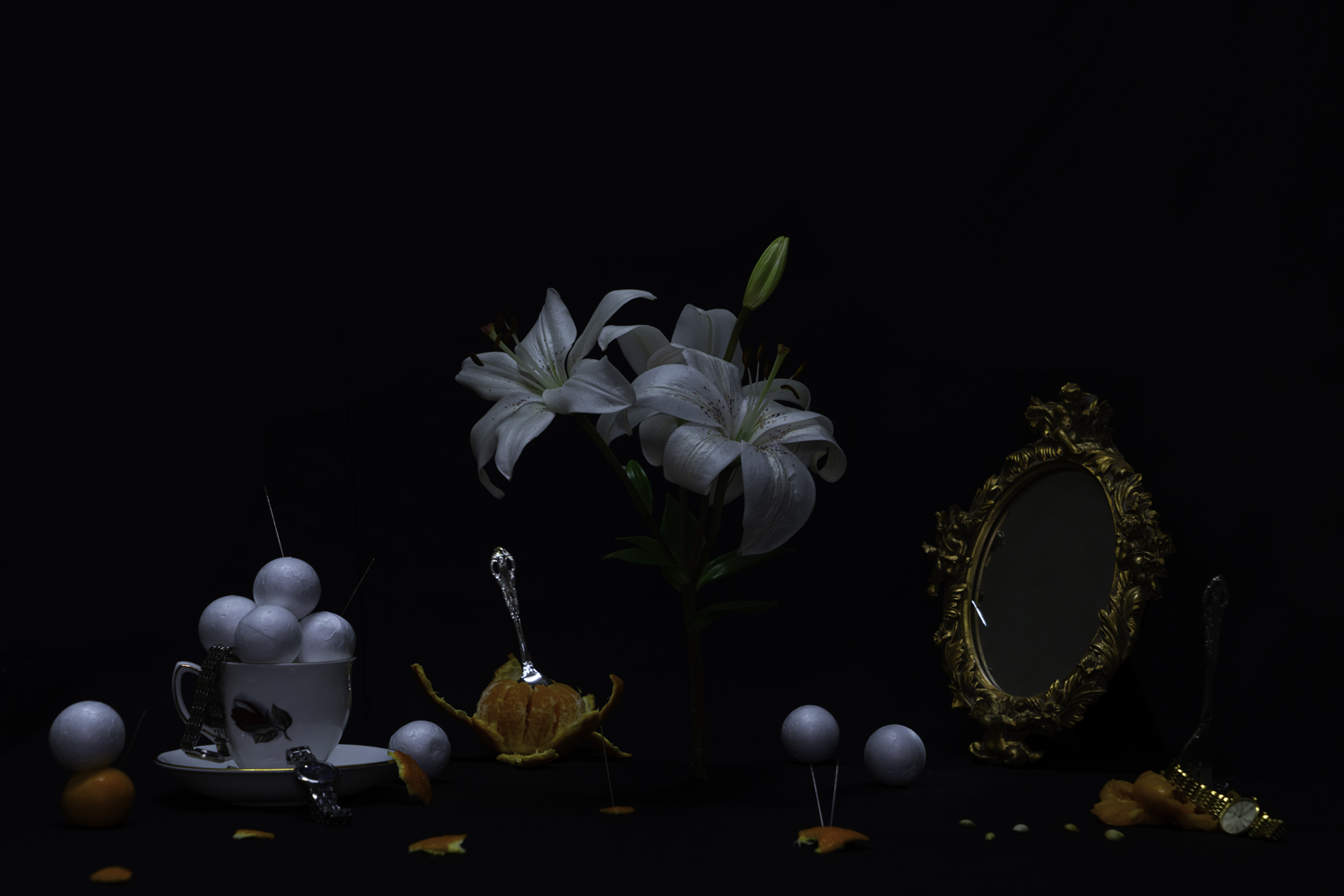 Dark digital photograph of table top with white lilies in vase with scattered food items, teacup, polystyrene balls and mirror.