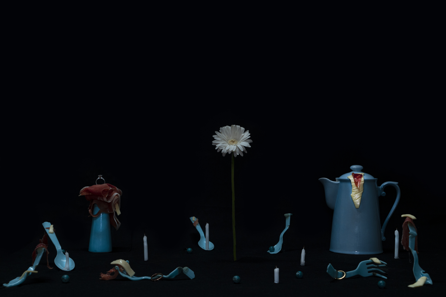 Dark digital image of daisy standing amongst scattered items of blue milk jugs, melted plastic cutlery, small white unlit candles and jewellery items. 