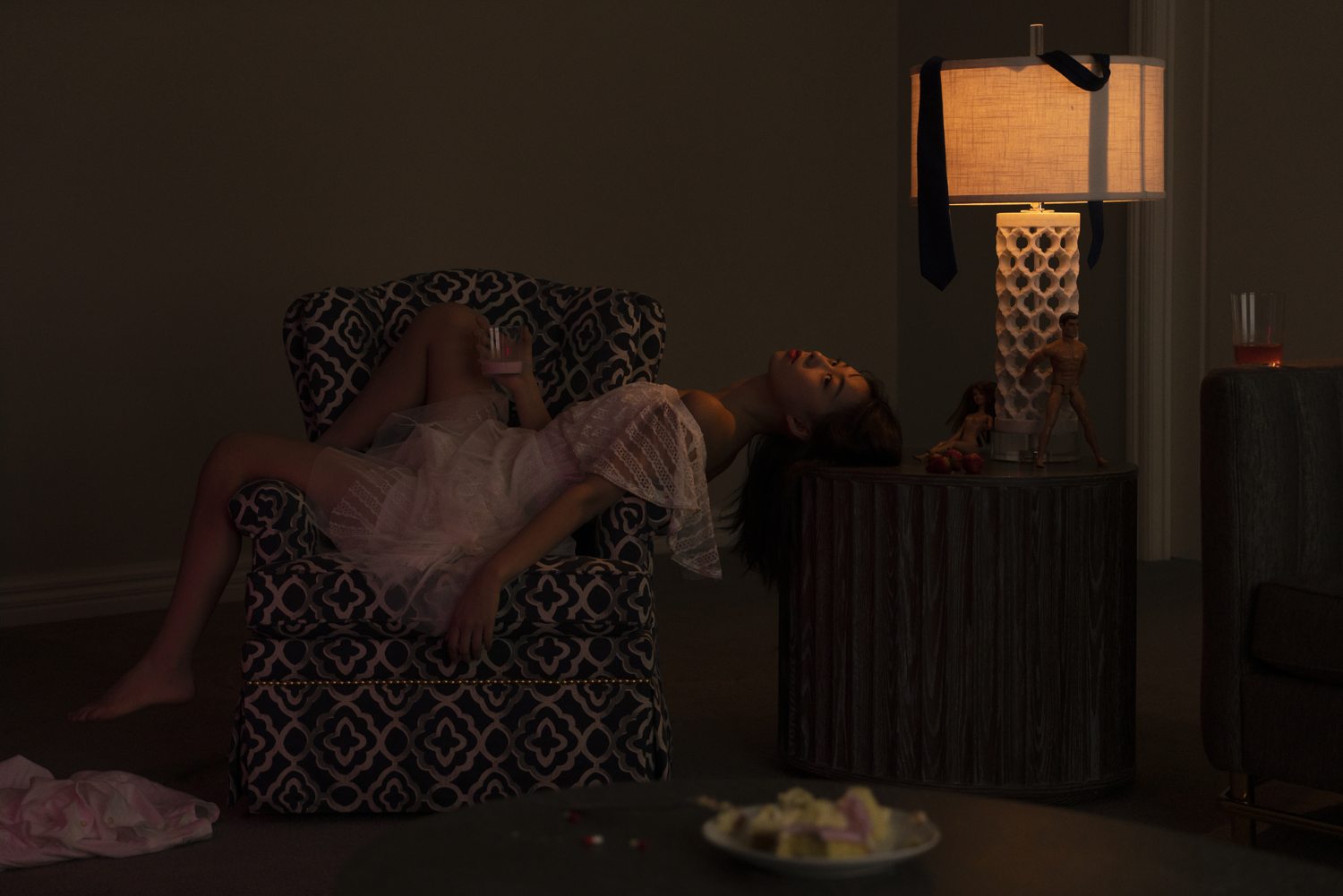 Digital photograph of woman in white dress lying across a small patterned lounge chair in dark lounge room.