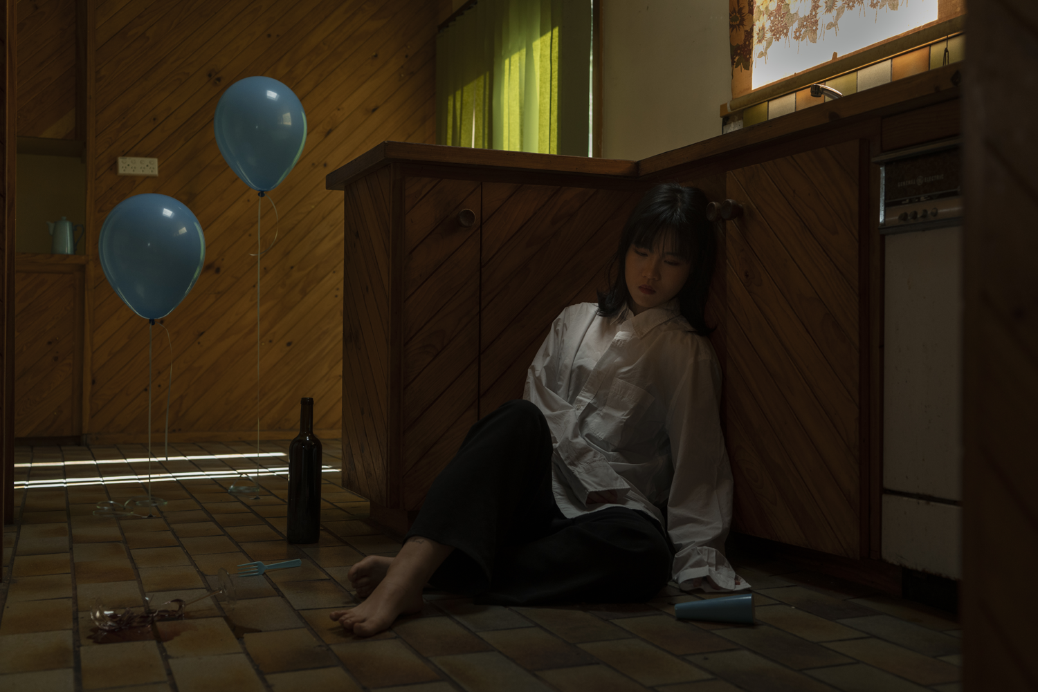 Dark digital image of woman sitting on tiled floor, leaning against a kitchen bench.  A wine bottle and two pale blue balloons sit on the floor beside her.  