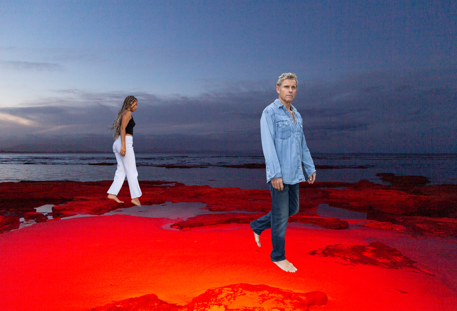 Colour image of woman and man standing on a rocky shoreline. Rock formations are cherry red. 