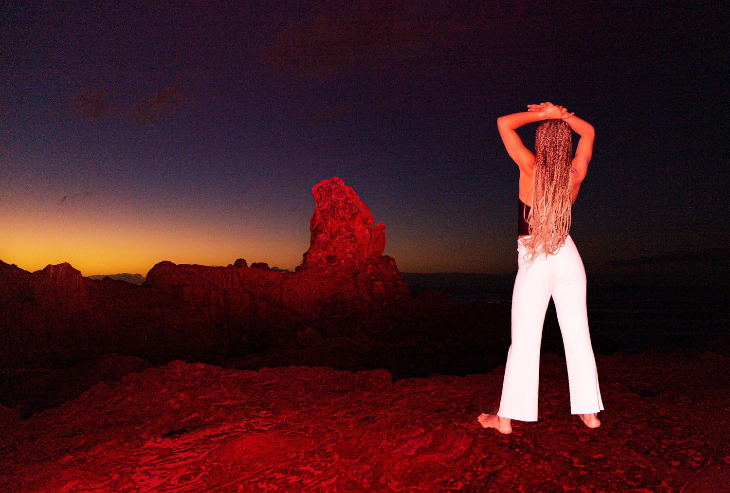 Colour image of woman standing in rocky landscape at dusk.  Woman stands with back to camera, facing the horizon.