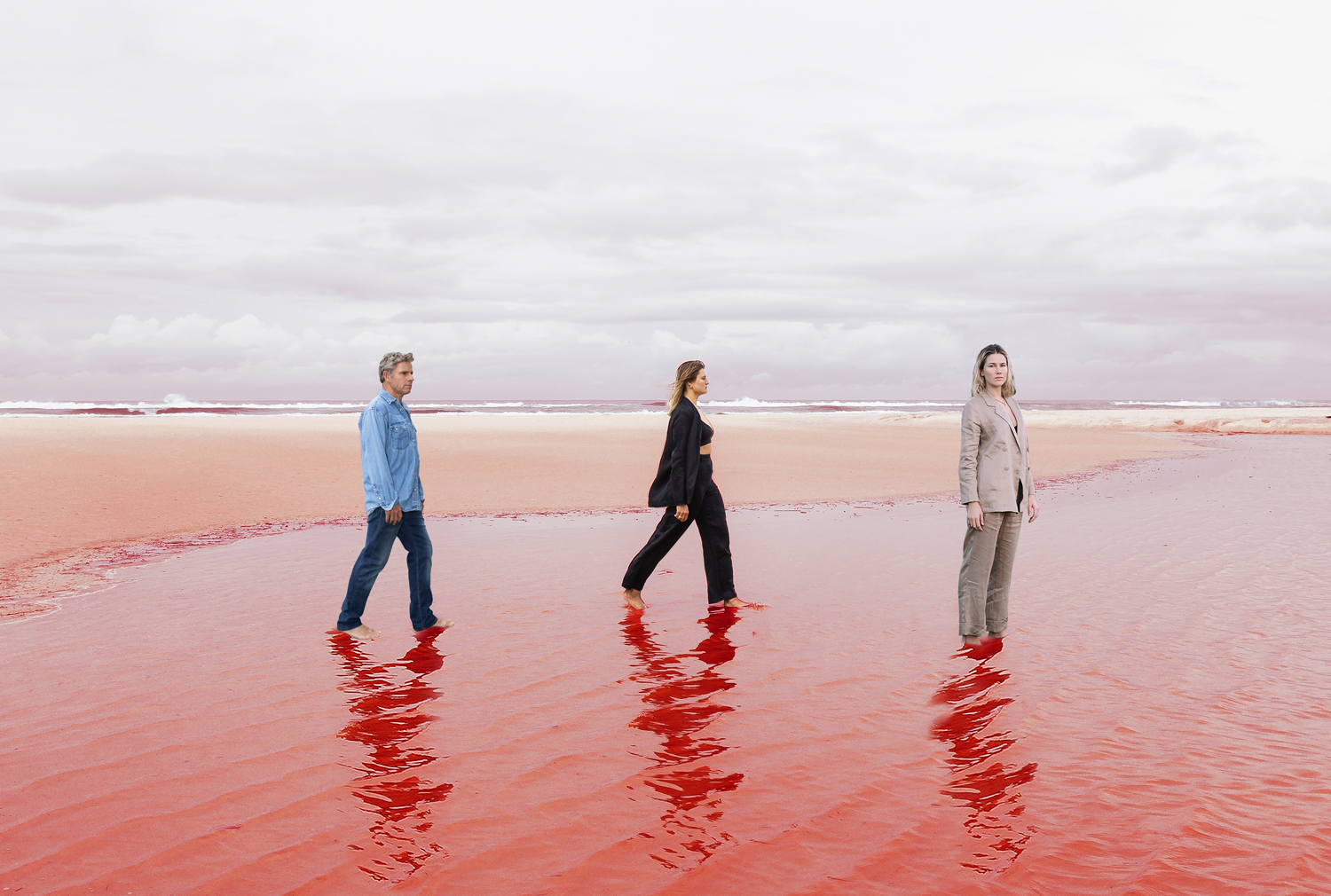 Colour image of three people walking consecutively in a line, on the bank of a red shoreline. 