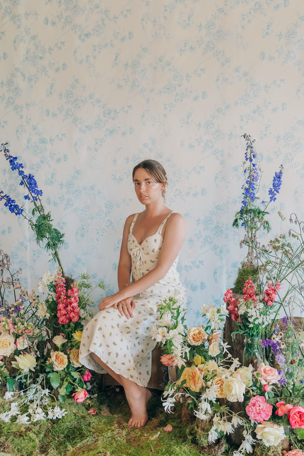 Colour photograph of a portrait of a young woman, in a floral dress sitting amongst greenery and an array of flowers. 