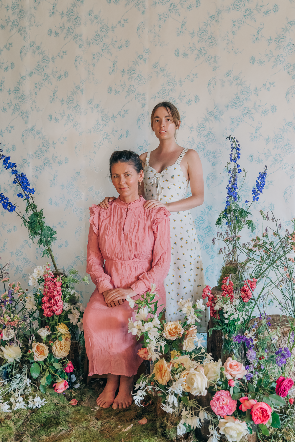 Colour photograph of a portrait of a young woman and older woman, sitting amongst greenery and an array of flowers. Young woman stands behind seated older woman.
