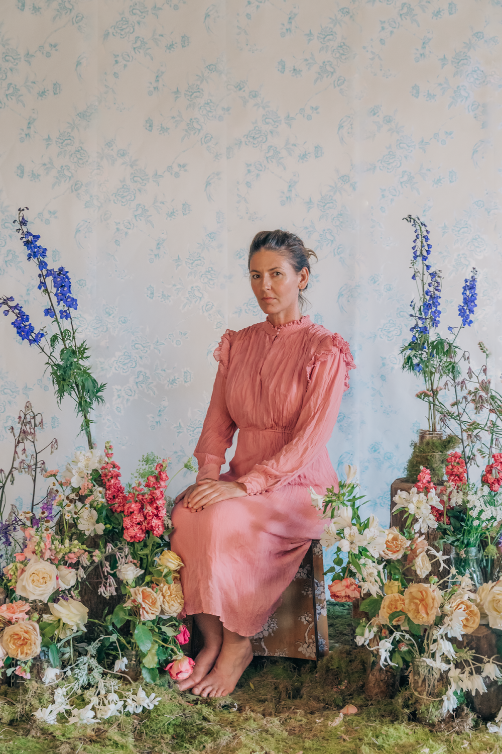 Colour photograph of a portrait of an older woman, in a salmon dress sitting amongst greenery and an array of flowers. 