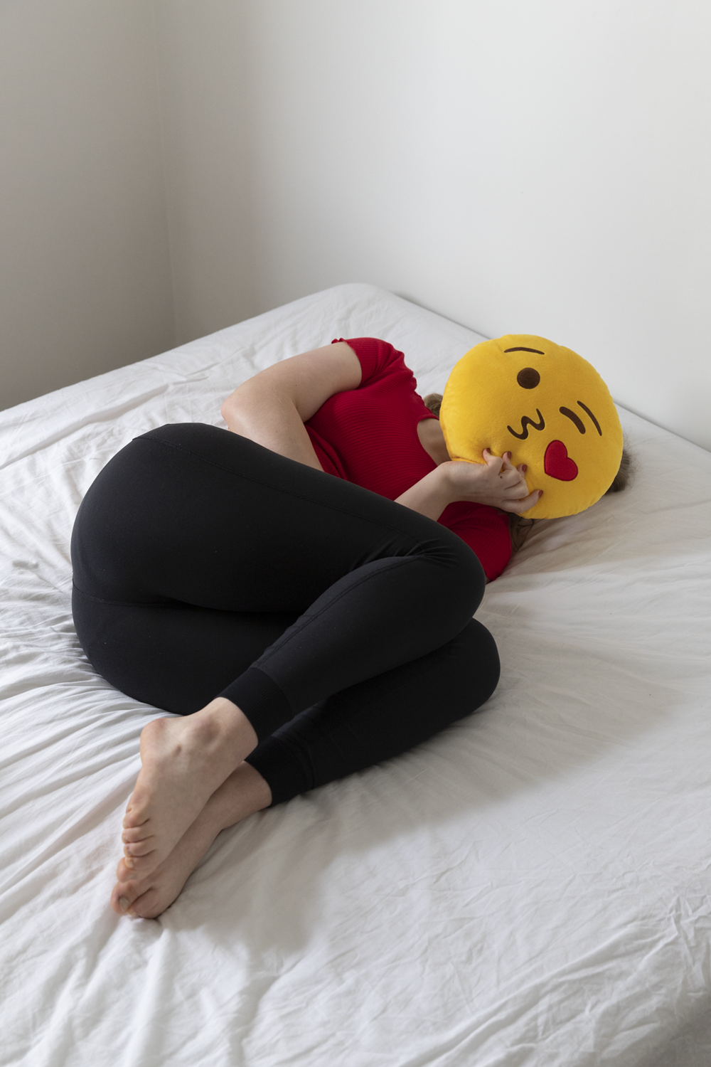 Colour photograph of female curled on empty bed with emoji pillow covering face.