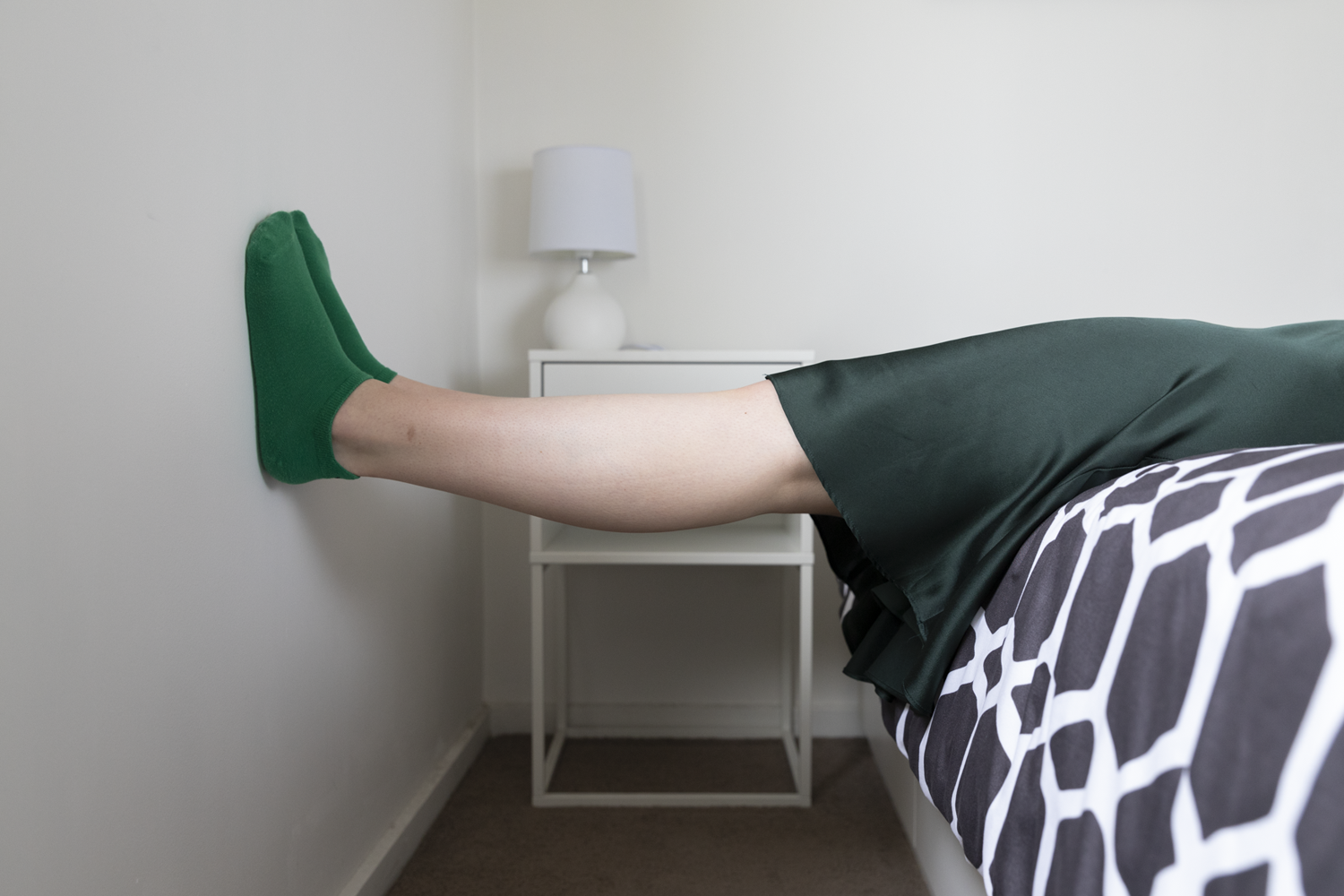 Colour photograph of legs in green skirt and green socks horizontally leaning on wall.