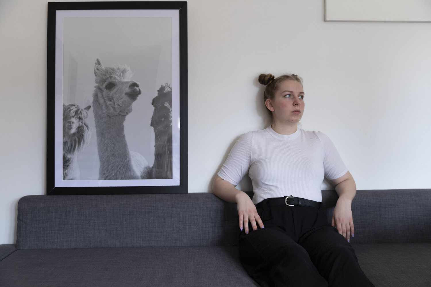 Colour photograph of female with space buns sitting next to framed image of llama, facing same direction.