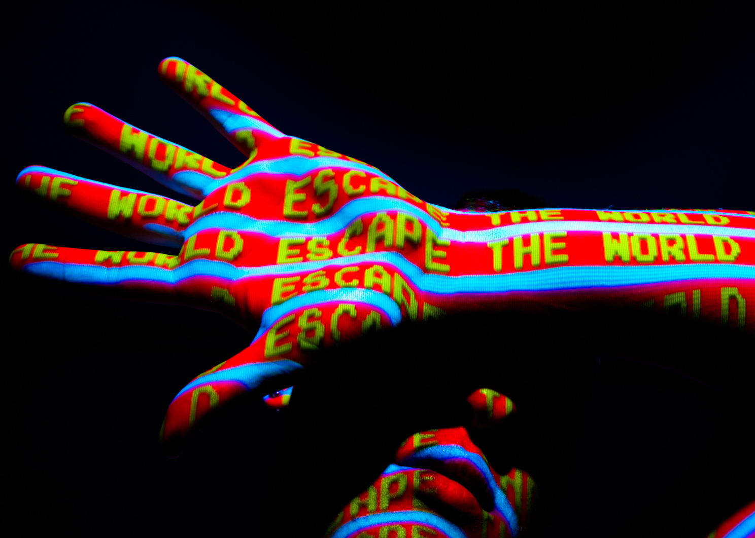 Colour image of a portrait of a man covering his face with his hand. Projected digital imagery covers his face. The words "escape the world" are displayed on his skin.