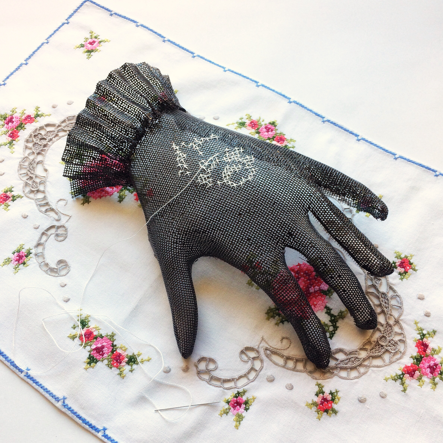 Enamelled copper mesh glove with stitching resting on found embroidered tray cloth
