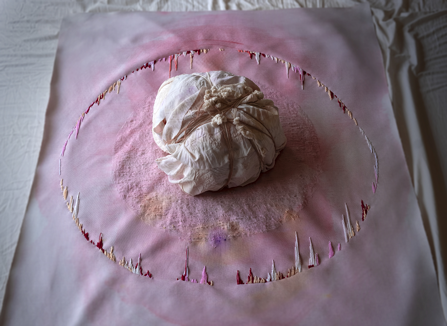 Documentation video of performer interacting with embroidered pink fabric; touching, laying underneath it. Performer is also wrapping yarn around a silk bundle dyed with flowers. 