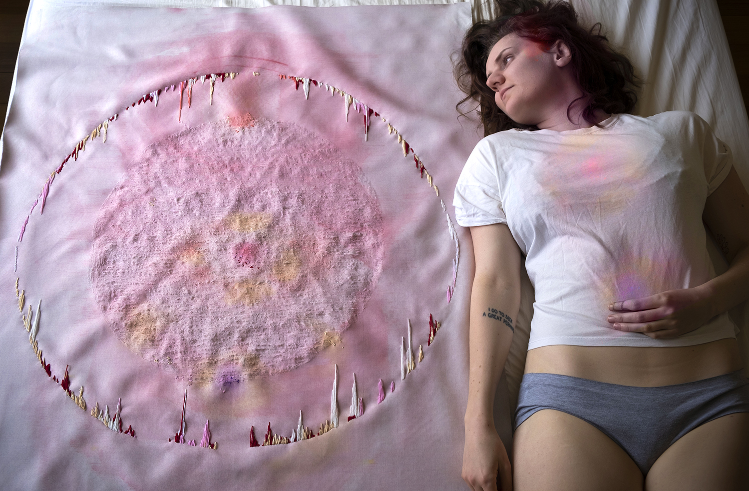 Linen fabric painted light pink. Roughed up circle in the middle of fabric with an embroidered circle around it. Performer laying next to fabric with repeated colors and patterns on their body.