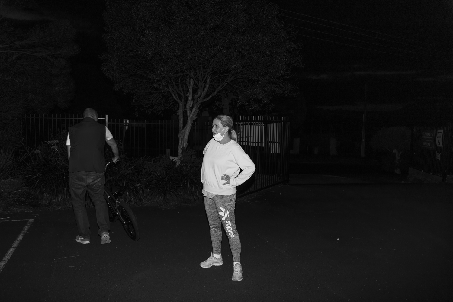 Black and white photograph of candid portrait of a woman wearing a mask alongside a man with bike in a car park at night.