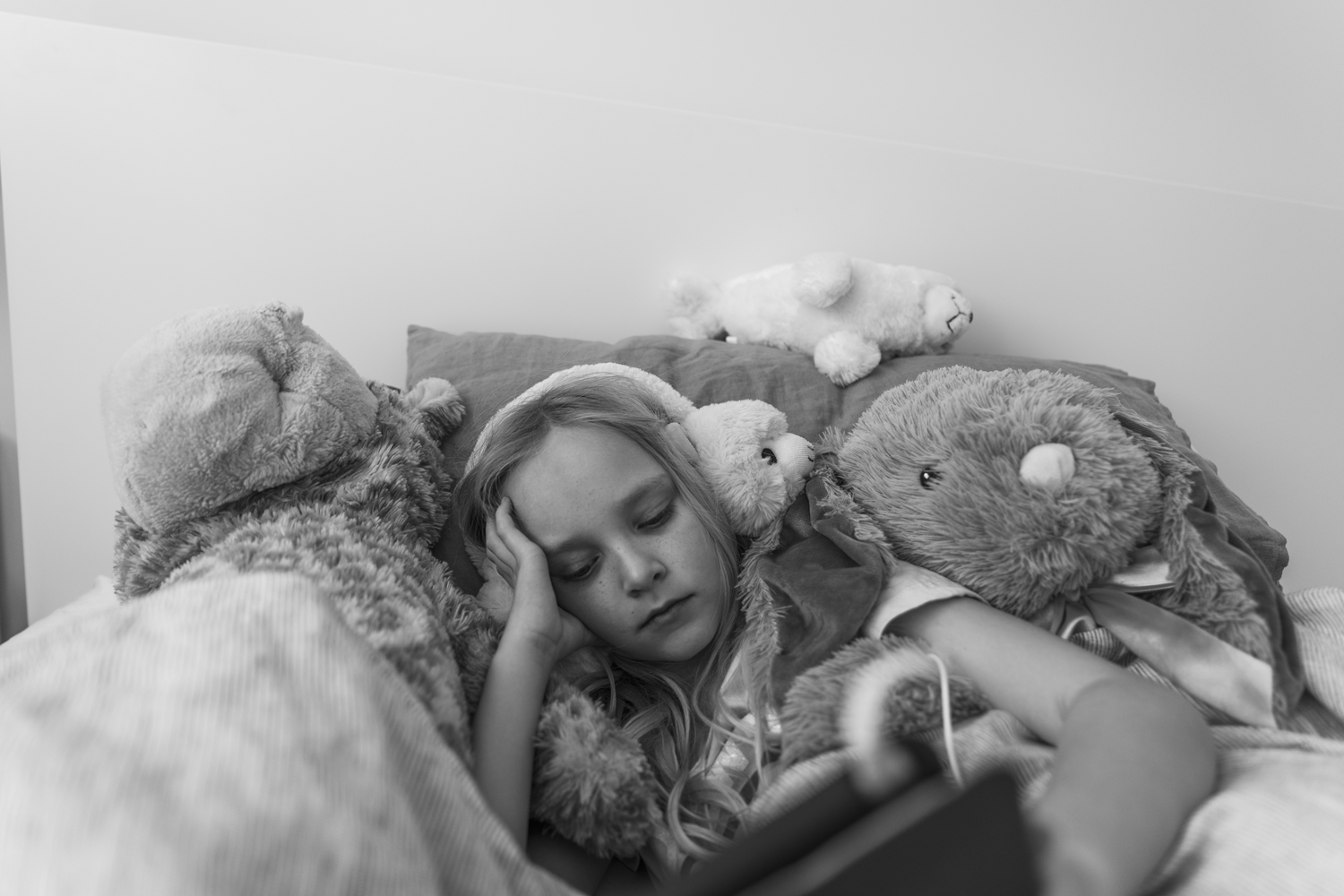Black and white photograph of a portrait of a girl lying amongst soft toys wearing headphones.