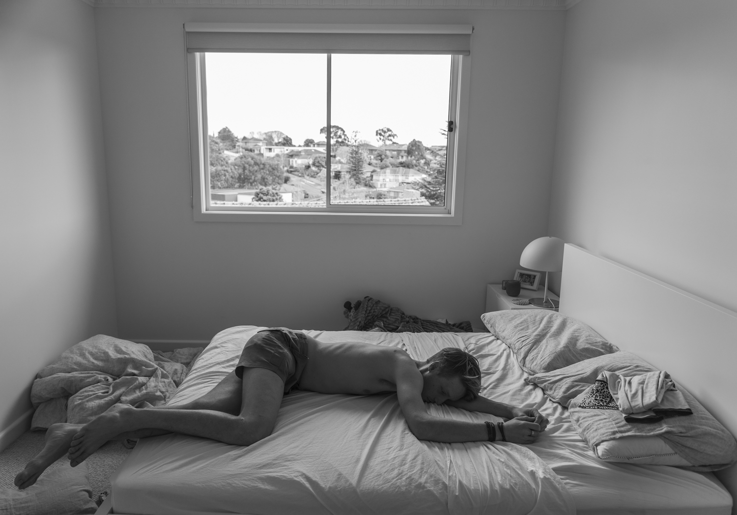 Black and white photograph of a bedroom interior with a teenage boy sleeping face down on a bed.