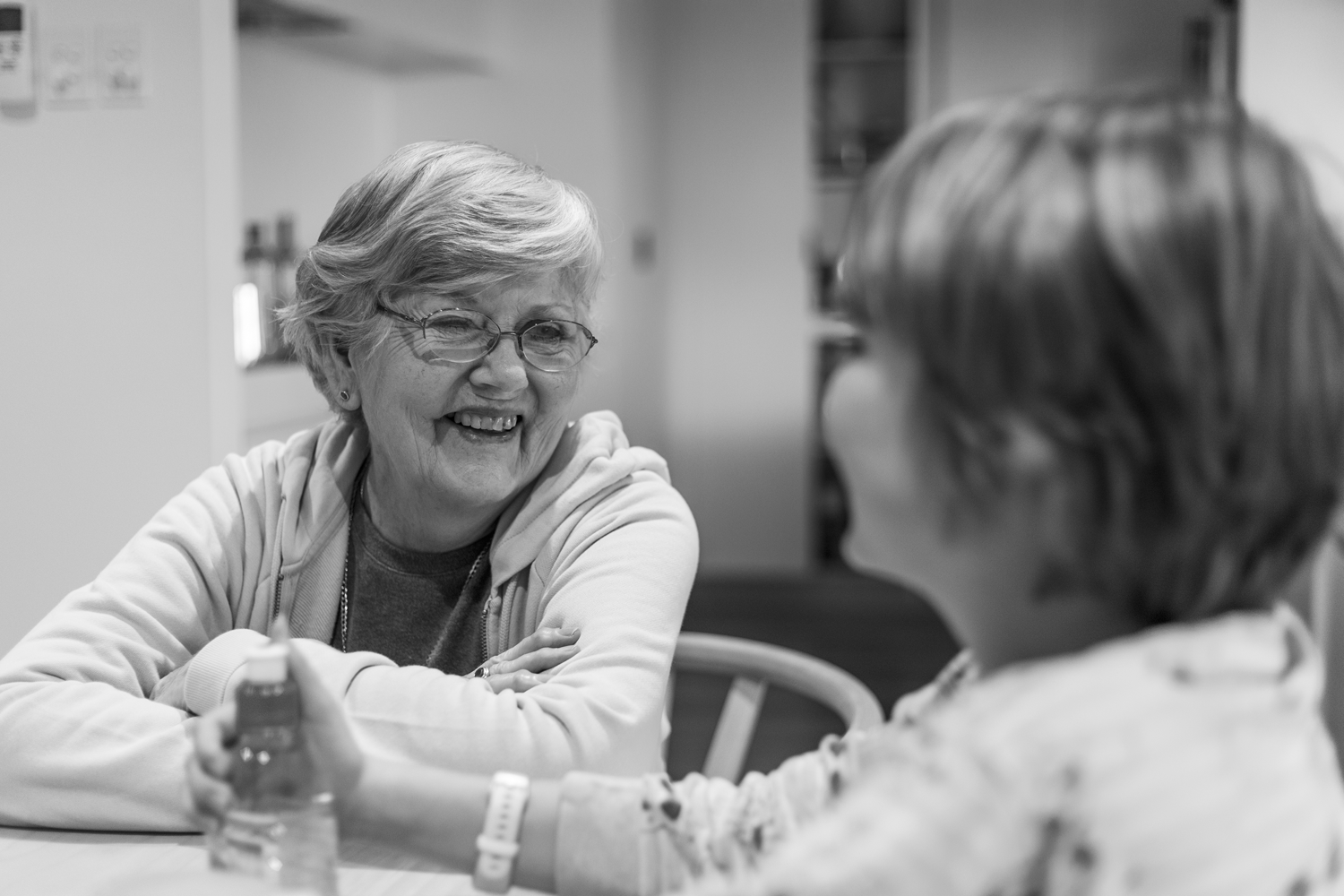Black and white photograph of a portrait of an elderly woman in laughing in conversation with another, in a domestic environment. 