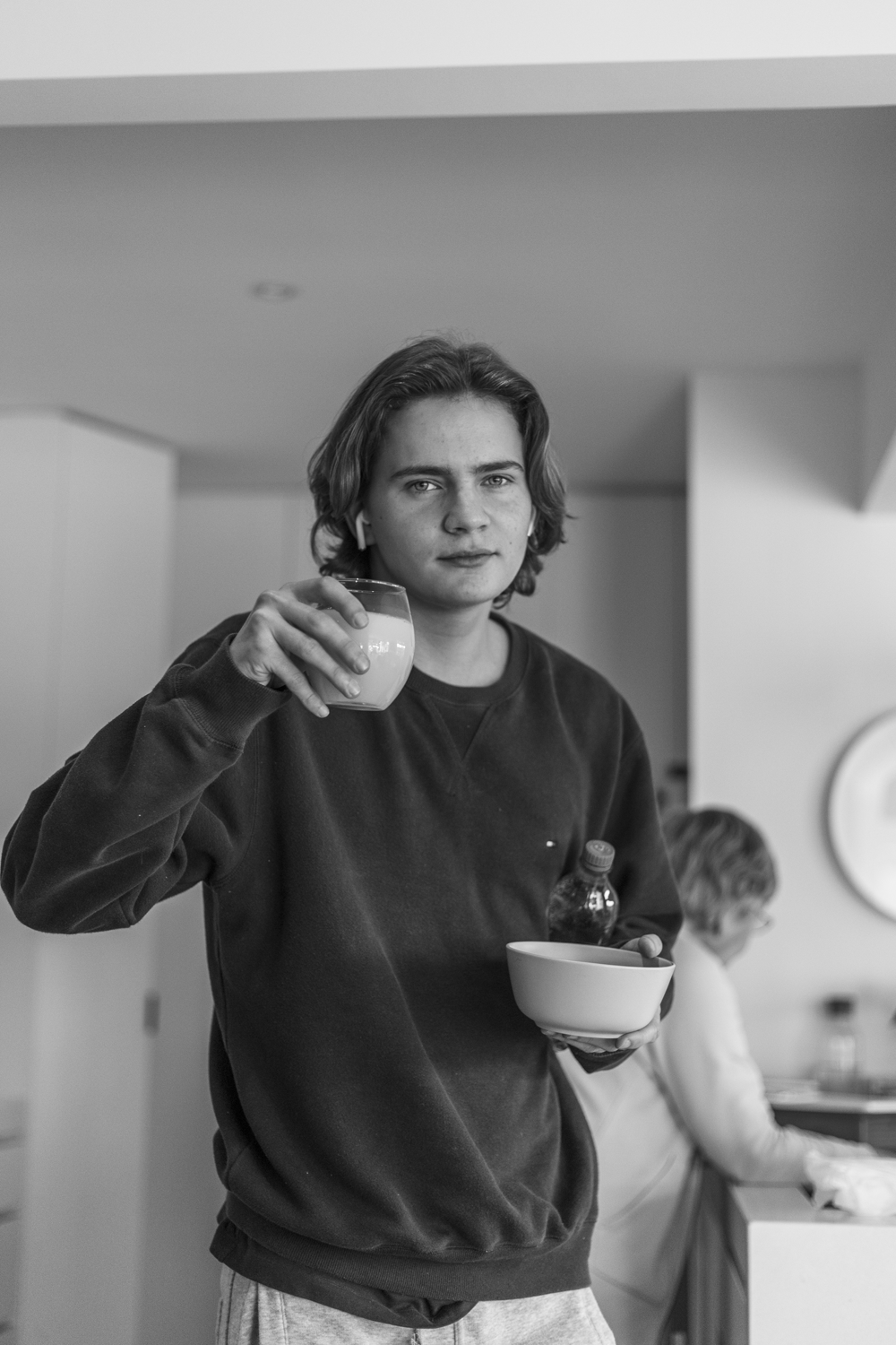 Black and white photograph of a teenage boy holding a glass and a bowl in a domestic kitchen environment. 