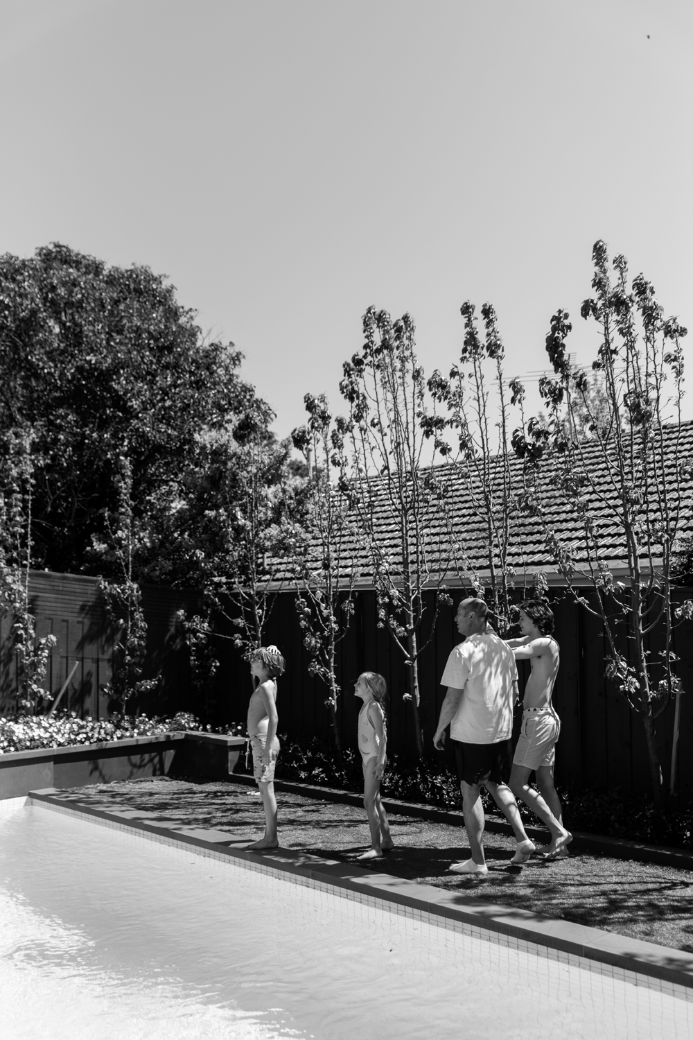 Black and white photograph of family standing poolside in the backyard of a domestic environment.