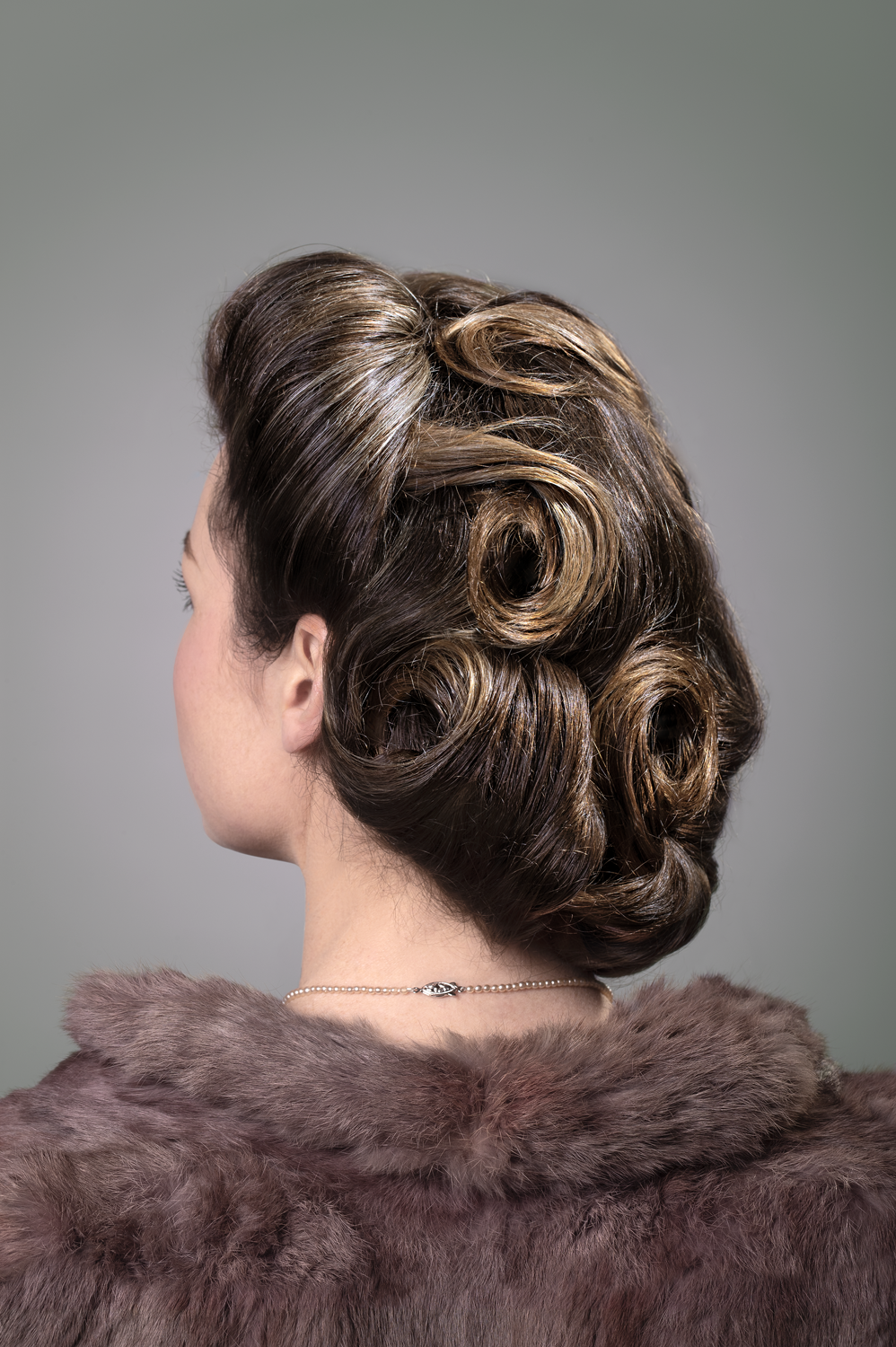 A feminine portrait from behind the head. 1940's hair style called Victory Roll. Pearls, fur jacket. Subject looking away into the distance face not visible. 