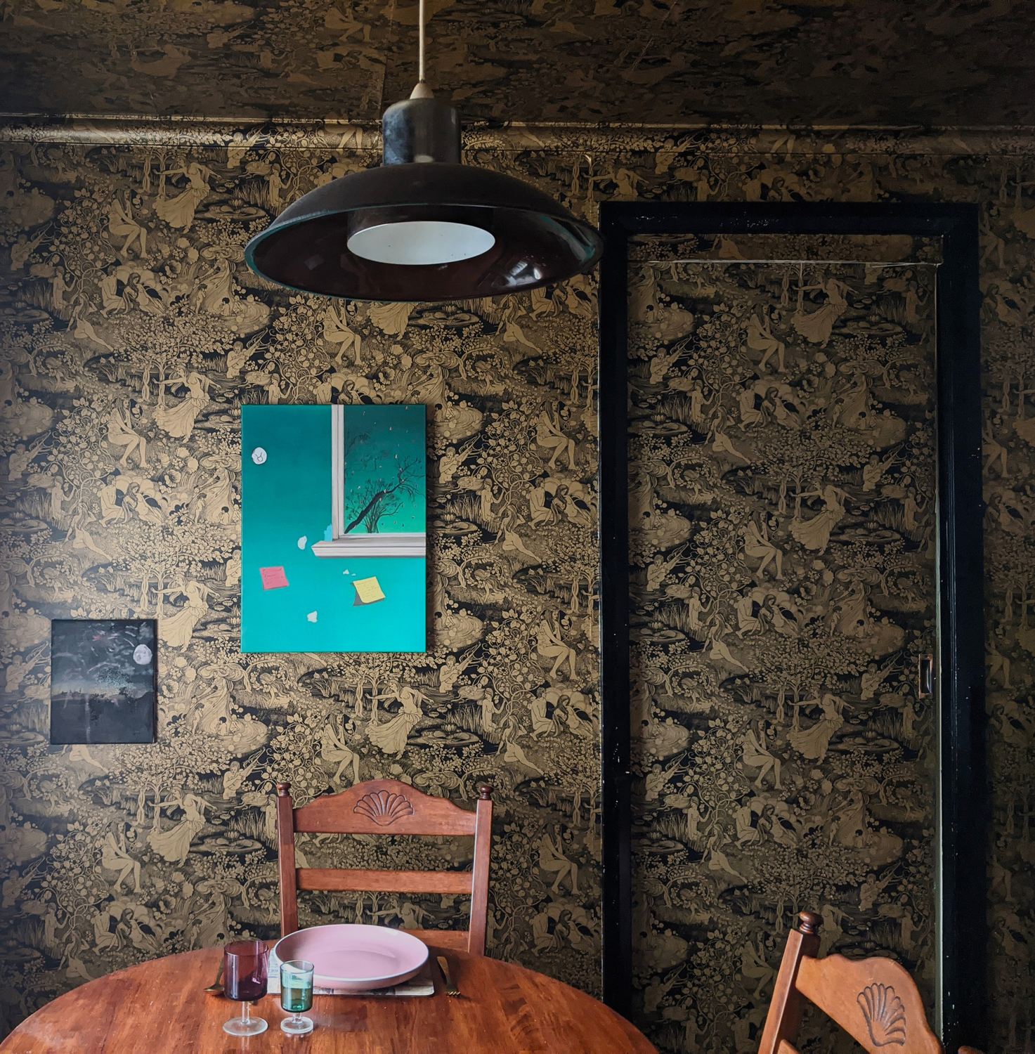 A family dining room. A gradient green painting hangs from the wall accompanied by a smaller painting of a landscape scene scribbled with fireworks.