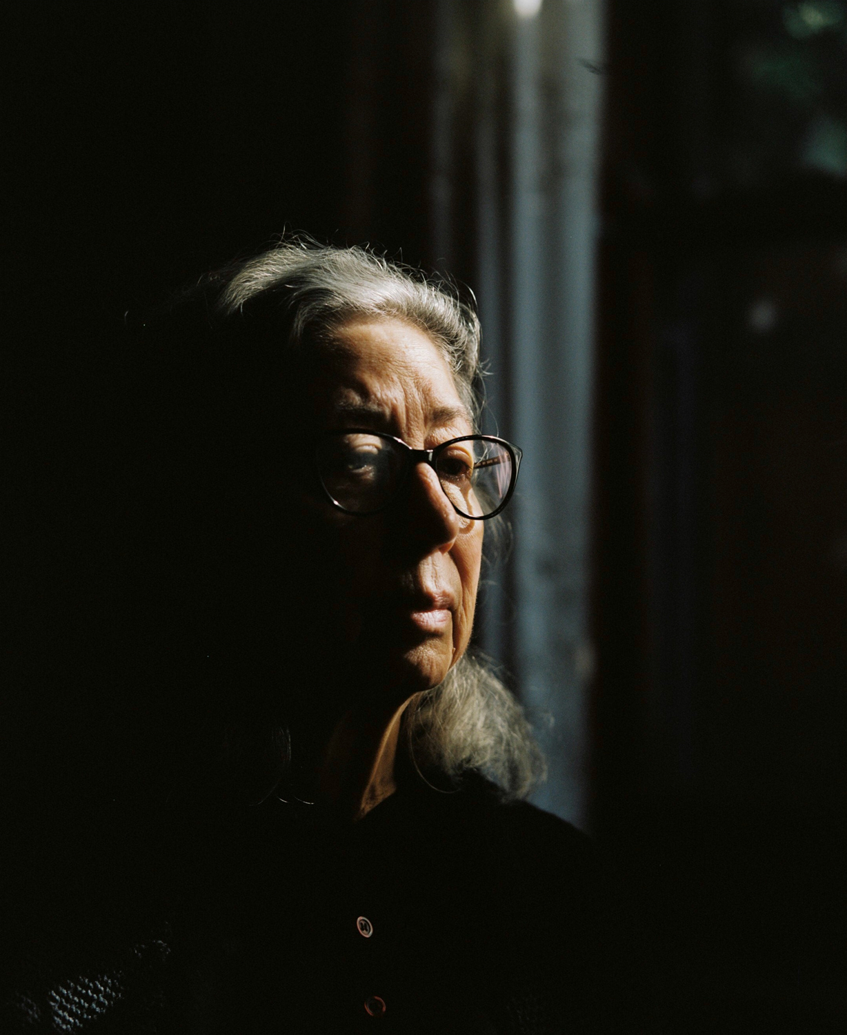 Colour photograph of a portrait of the artist's mother. Woman sits by window in dappled light.