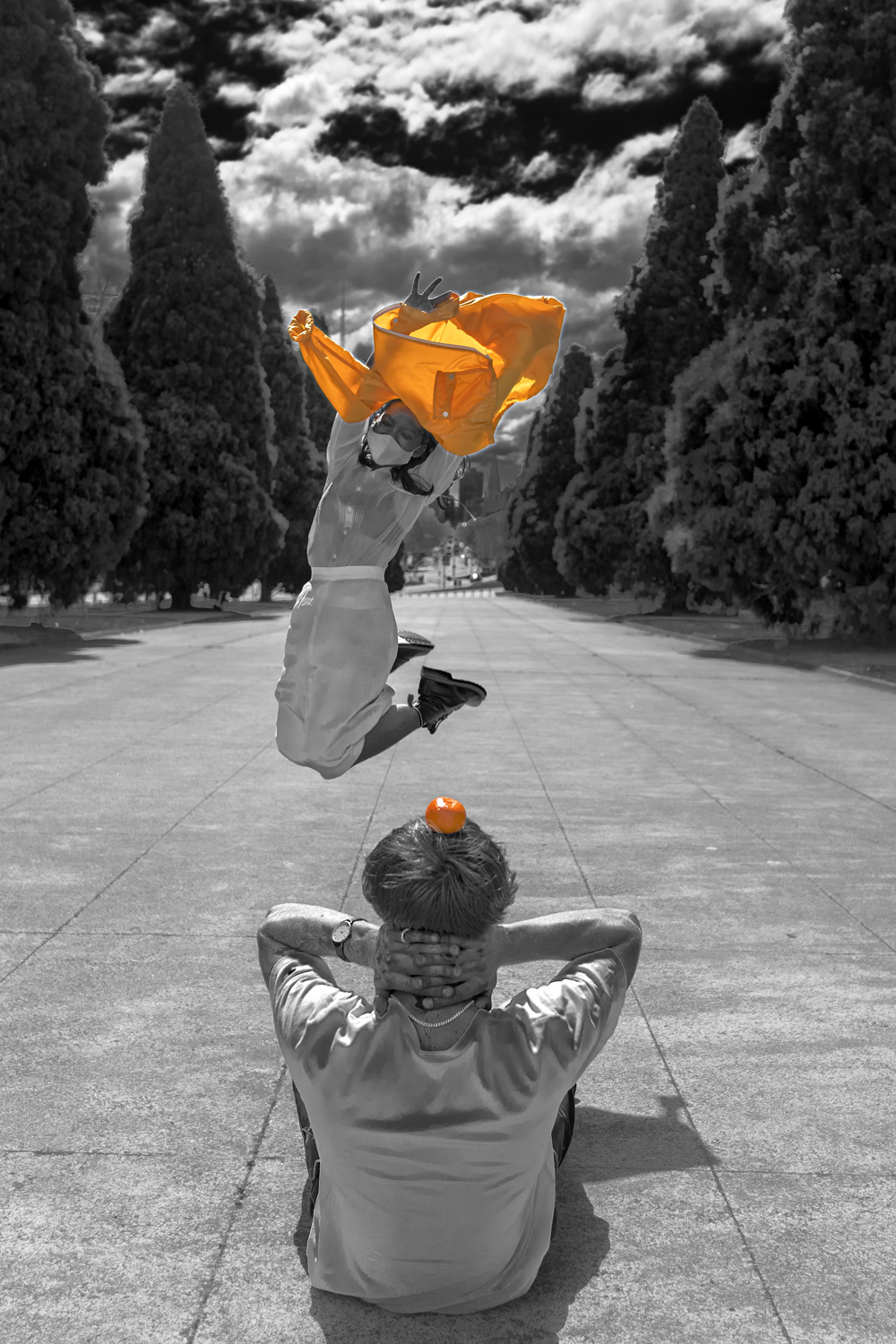 Black and white photograph with yellow details depicting a man sitting on the ground  with an orange on his head and a woman jumping the air with her smock above her head.