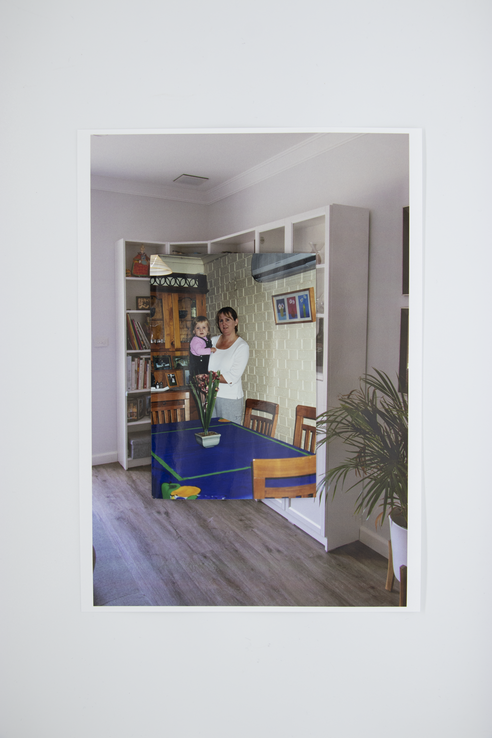 Coloured photograph depicting mother holding child in domestic dining room beside a kitchen table. Photo is bordered by a secondary image depicting a corner of an empty room with white bookshelves.
