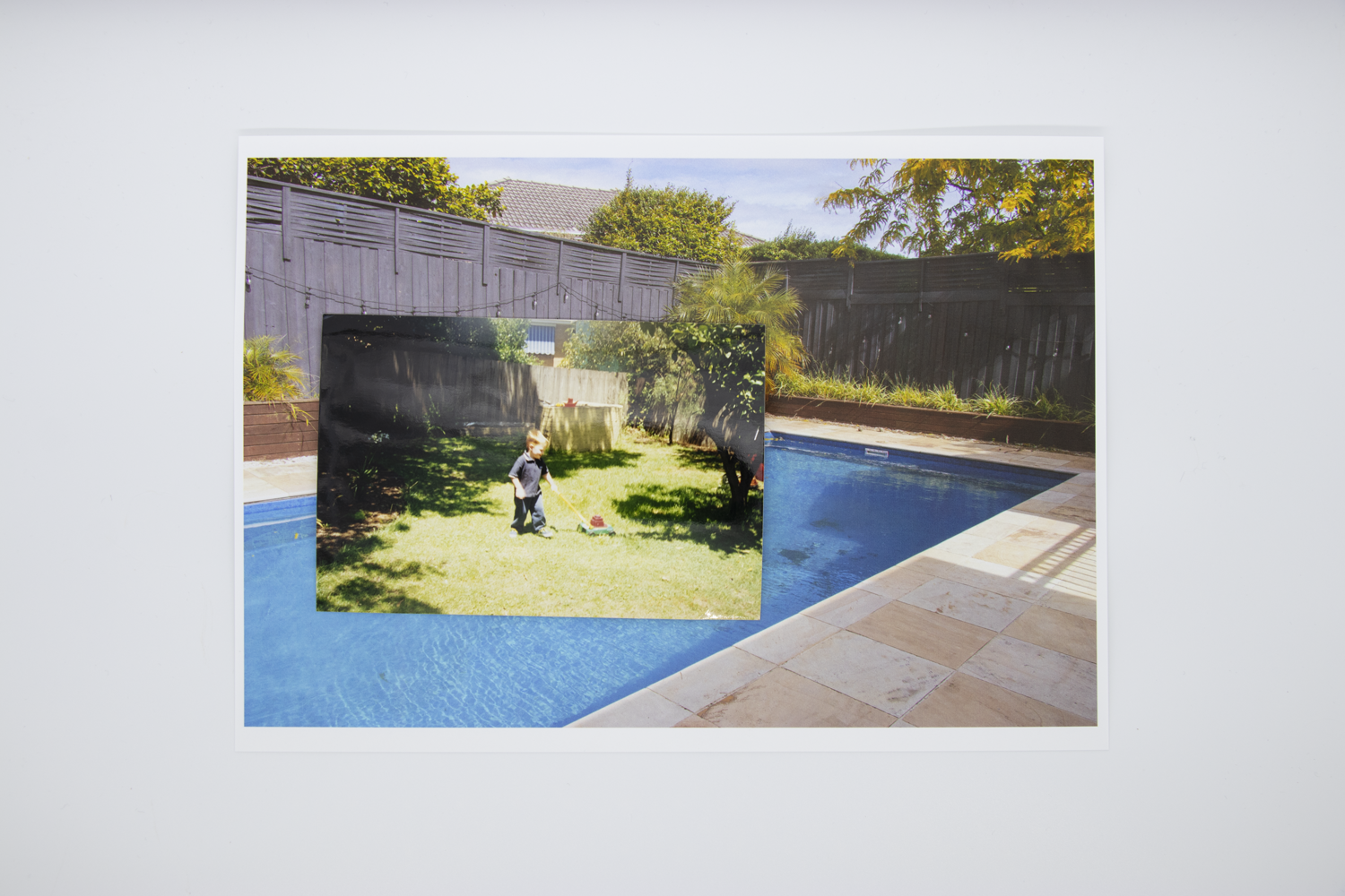 Coloured photograph depicting child pushing toy lawnmower in suburban backyard. Photo is bordered by secondary image of blue pool in suburban backyard.