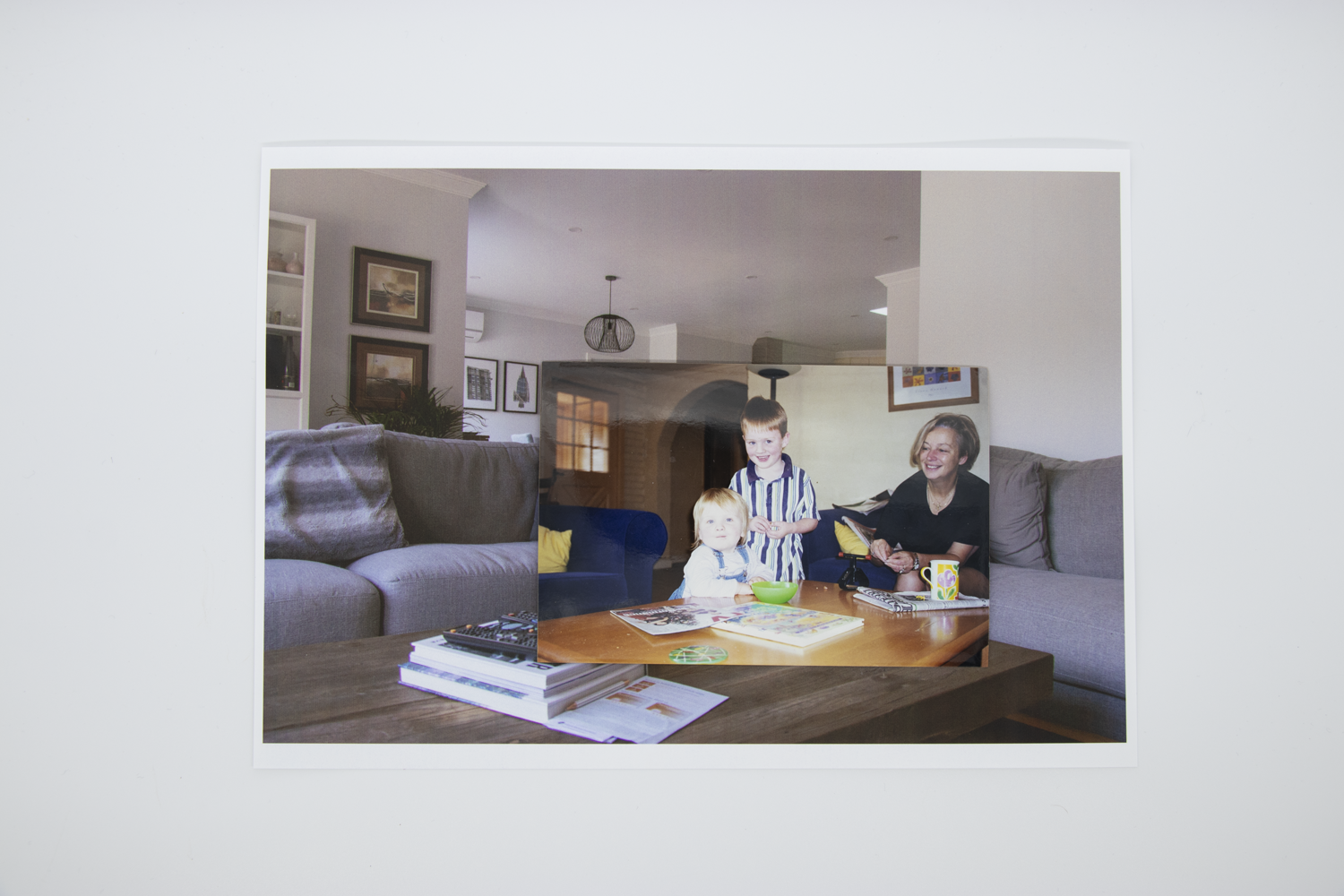 Coloured photograph depicting two children and mother in a domestic lounge room. Photo is bordered by a secondary image of an empty domestic lounge room with grey couches.