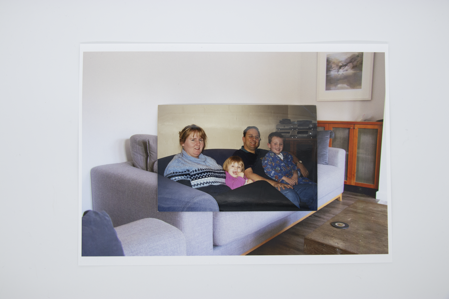 Coloured photograph depicting two children and mother sitting on a couch in a domestic lounge room. Photo is bordered by a secondary image of an empty domestic lounge room with grey couches.