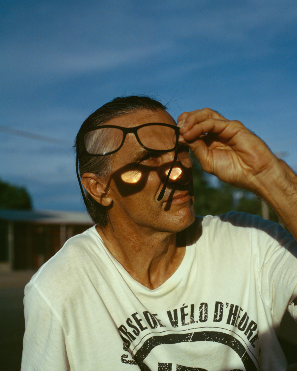 Colour photograph depicting man holding up glasses toward sun. Light refracts through glass onto face.