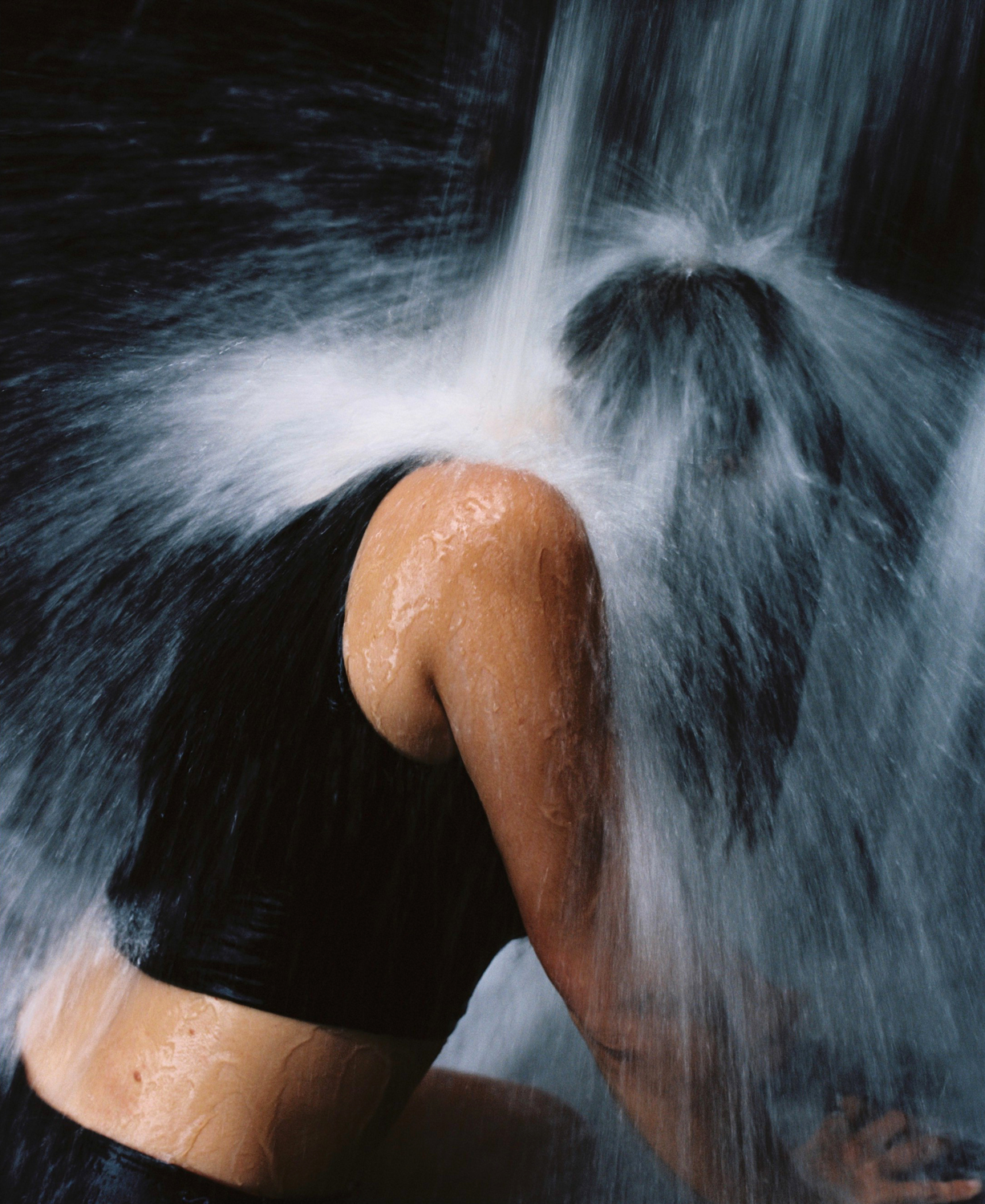 Colour photograph depicting girl with head under running waterfall. 